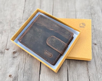 May include: A brown leather wallet with a snap closure, presented in a yellow box. The wallet has a distressed finish and the word "MUDD" is embossed on the front. The box has an RFID symbol.