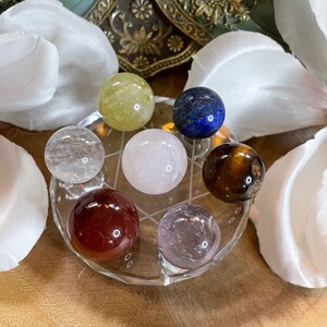 May include: Seven crystal spheres of various colors, including clear, yellow, pink, blue, brown, red, and purple, are arranged in a circular pattern on a clear crystal stand.