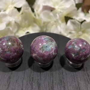 May include: Three polished gemstone spheres with a mix of colors, including deep purple, teal, and dark green. The spheres are displayed on a black stand, with white flowers in the background. These decorative spheres are ideal for home decor.