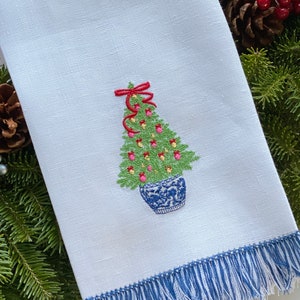 Pu&ograve; includere: Un asciugamano da cucina bianco con un bordo a frangia blu presenta un ricamo di un albero di Natale in vaso con ornamenti rossi, fiori rosa e un fiocco rosso.