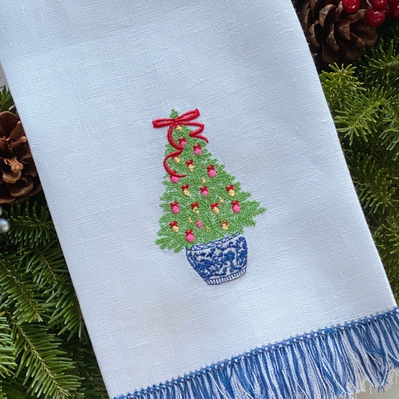 Pu&ograve; includere: Un asciugamano da cucina bianco con un bordo a frangia blu presenta un ricamo di un albero di Natale in vaso con ornamenti rossi, fiori rosa e un fiocco rosso.