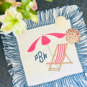 May include: A white linen napkin with blue fringe, embroidered with a pink and white beach umbrella, a striped beach chair, and the monogram "PBW".