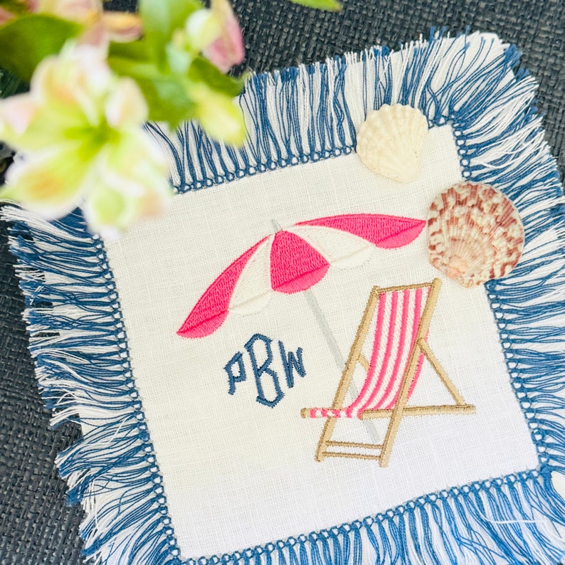 May include: A white linen napkin with blue fringe, embroidered with a pink and white beach umbrella, a striped beach chair, and the monogram "PBW".