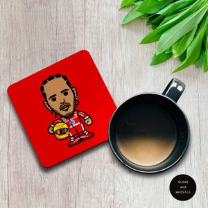 May include: Red square coaster with a cartoon illustration of a Formula 1 driver wearing a red racing suit and helmet. The driver is holding a yellow helmet in his left hand. The coaster is on a wooden surface.