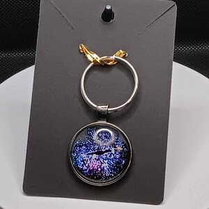 May include: A silver key chain with a round, blue, purple, and black galaxy-themed glass charm. The charm is attached to a silver ring.