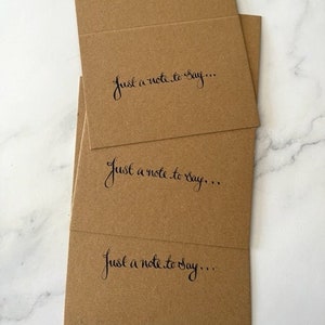 May include: Kraft paper greeting cards with the handwritten phrase "Just a note to say..." in black script. The cards are stacked, with the top card slightly open, revealing the inside. The cards are a warm brown colour.