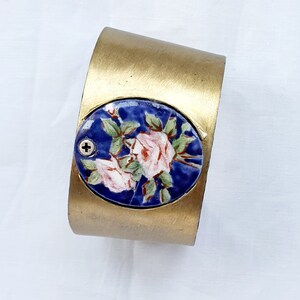 Könnte beinhalten: Ein goldfarbenes Metall-Armband mit einer ovalen, blauen und weißen Keramik-Einlage mit einem floralen Design. Die Einlage zeigt zwei rosa Blüten mit grünen Blättern und einem blauen Hintergrund.