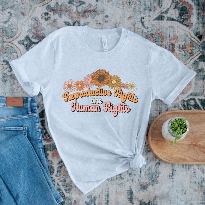 Puede incluir: Una camiseta gris claro con un estampado gr&aacute;fico de flores y el texto "Reproductive Rights are Human Rights" en una fuente retro.