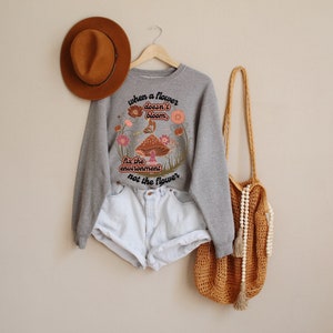 K&ouml;nnte beinhalten: Grauer Pullover mit einem Blumenmuster und dem Text "Wenn eine Blume nicht bl&uuml;ht, repariere die Umwelt, nicht die Blume".