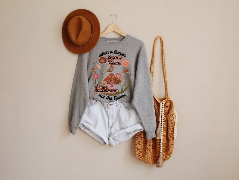 K&ouml;nnte beinhalten: Grauer Pullover mit einem Blumenmuster und dem Text "Wenn eine Blume nicht bl&uuml;ht, repariere die Umwelt, nicht die Blume".
