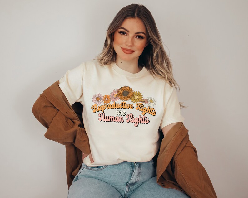 Puede incluir: Una camiseta blanca con un dise&ntilde;o floral y el texto "Reproductive Rights are Human Rights" en una fuente retro.