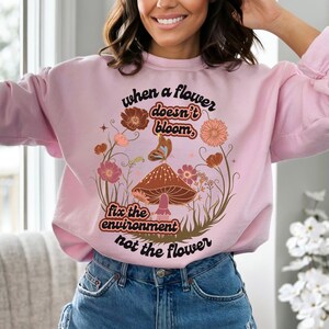 K&ouml;nnte beinhalten: Rosafarbener Pullover mit einem Aufdruck eines Pilzes und Blumen. Der Text auf dem Pullover lautet "When a flower doesn't bloom, fix the environment not the flower."