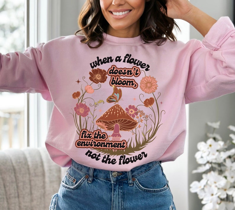 K&ouml;nnte beinhalten: Rosafarbener Pullover mit einem Aufdruck eines Pilzes und Blumen. Der Text auf dem Pullover lautet "When a flower doesn't bloom, fix the environment not the flower."