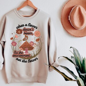 K&ouml;nnte beinhalten: Ein beiger Pullover mit einem Blumendesign und dem Text "when a flower doesn't bloom, fix the environment not the flower".