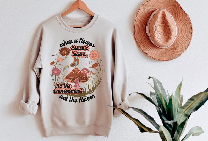 K&ouml;nnte beinhalten: Ein beiger Pullover mit einem Blumendesign und dem Text "when a flower doesn't bloom, fix the environment not the flower".