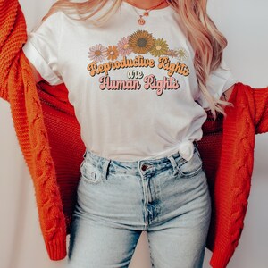 Puede incluir: Una camiseta blanca con un dise&ntilde;o gr&aacute;fico de flores y el texto "Reproductive Rights are Human Rights" en una fuente retro.