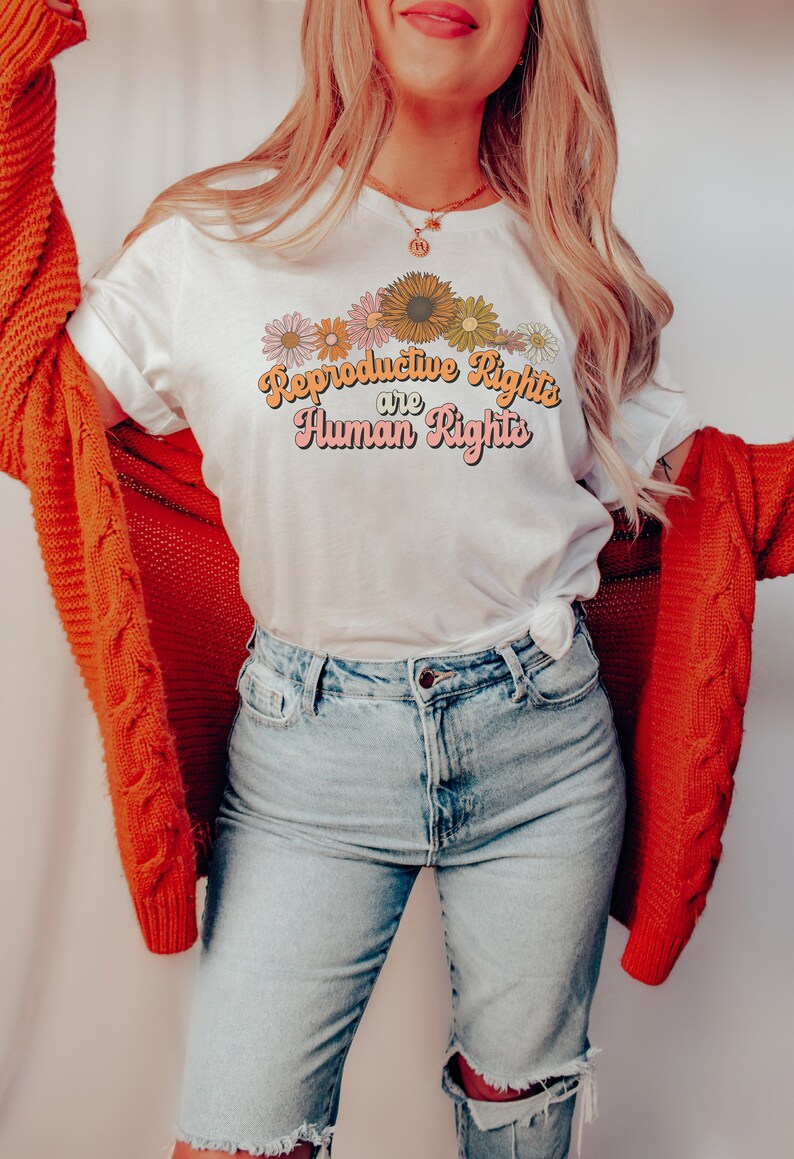 Puede incluir: Una camiseta blanca con un dise&ntilde;o gr&aacute;fico de flores y el texto "Reproductive Rights are Human Rights" en una fuente retro.