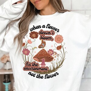 K&ouml;nnte beinhalten: Wei&szlig;es Sweatshirt mit einem grafischen Design aus einem Pilz, Blumen und Schmetterlingen. Der Text lautet "Wenn eine Blume nicht bl&uuml;ht, repariere die Umwelt, nicht die Blume."