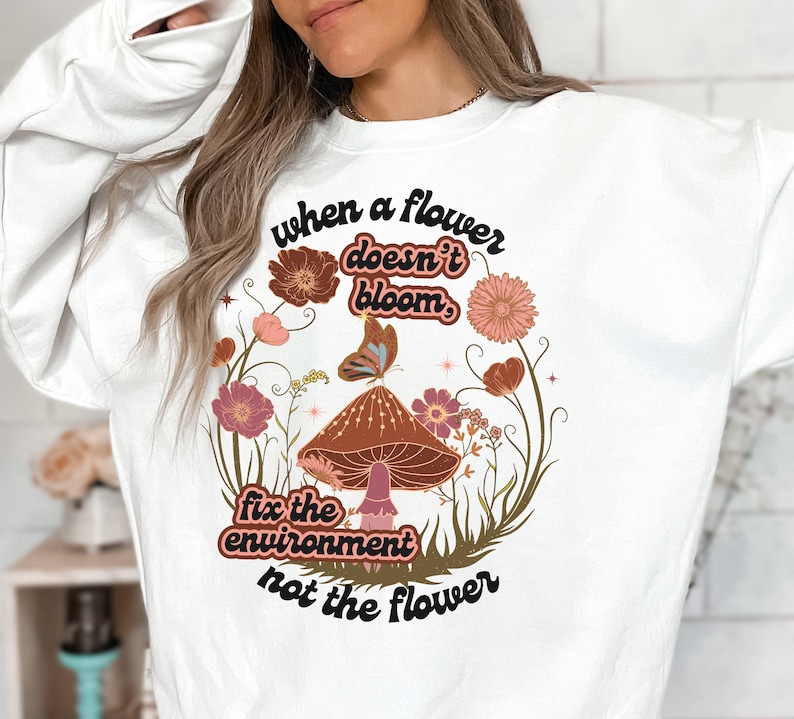 K&ouml;nnte beinhalten: Wei&szlig;es Sweatshirt mit einem grafischen Design aus einem Pilz, Blumen und Schmetterlingen. Der Text lautet "Wenn eine Blume nicht bl&uuml;ht, repariere die Umwelt, nicht die Blume."