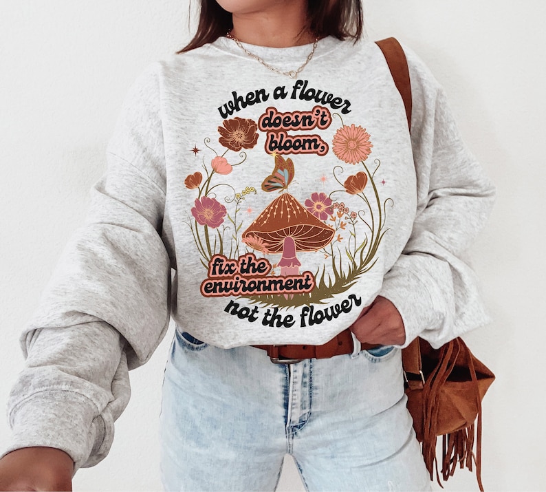 K&ouml;nnte beinhalten: Ein graues Sweatshirt mit einem grafischen Druck eines Pilzes und Blumen. Der Text auf dem Sweatshirt lautet "when a flower doesn't bloom, fix the environment not the flower".