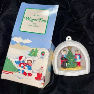 Puede incluir: Un adorno de árbol de Navidad de plástico blanco con forma de iglú con un pequeño árbol de Navidad y tres figuras en su interior. La caja dice "Avon Winter Fun Tree Ornament".