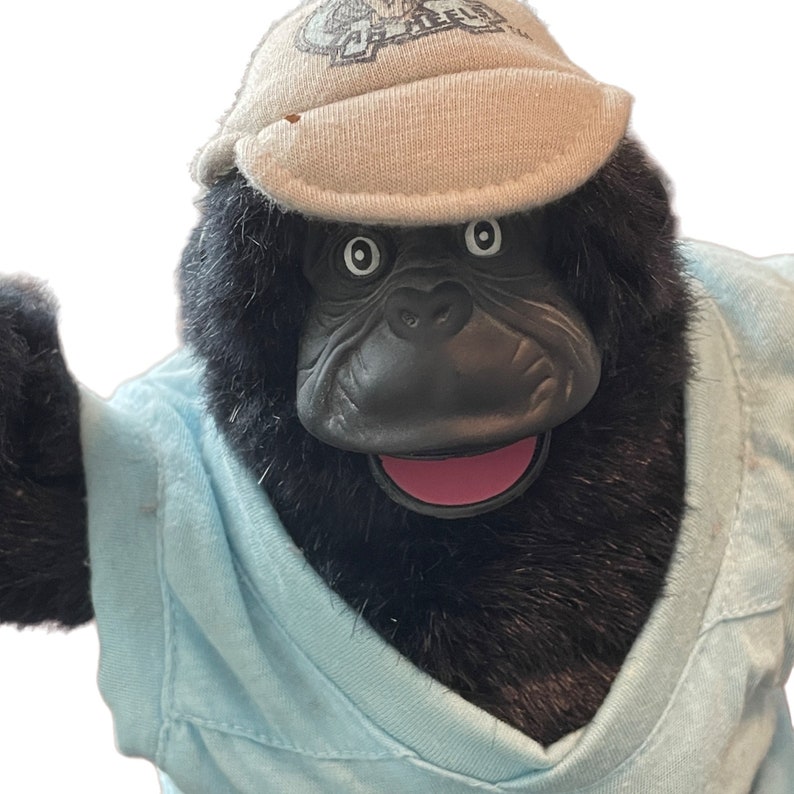 Op de afbeelding: Een zwart gorilla knuffeldier dat een lichtblauw shirt en een beige baseballpet draagt met de tekst "Tell"