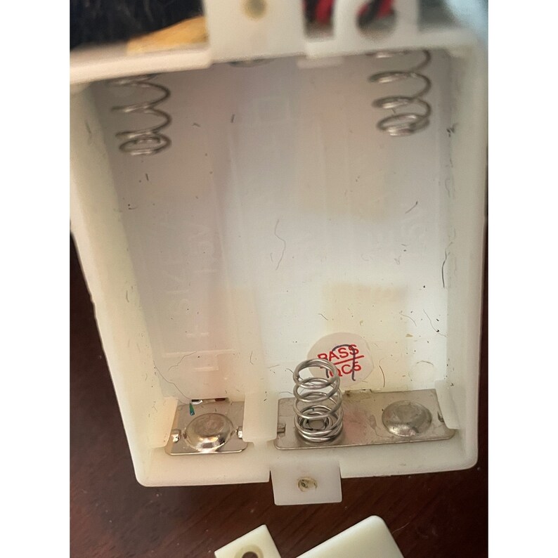 Op de afbeelding: Close-up van een wit plastic batterijvak met twee veerbelaste aansluitingen en een rode sticker met de tekst "Bass Inc."