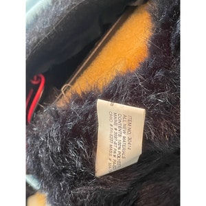 Op de afbeelding: Een zwarte bontmuts met een wit label eraan bevestigd. Het label heeft de volgende tekst: "ITEM NO. 30414 ALL NEW MATERIALS CONTENTS: 100% POLYESTER MAINE #3697-ST PA # PA-52 OHIO #PA-5207 MASS # MA-13"