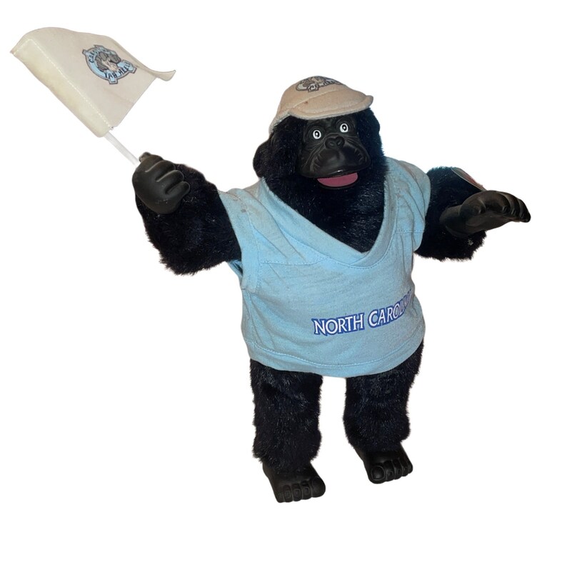 Op de afbeelding: Een zwarte pluche gorilla die een blauw shirt draagt met de tekst "NORTH CAROLINA" en een beige baseballpet. De gorilla houdt een witte vlag vast met de tekst "TAR HEELS" in blauw.