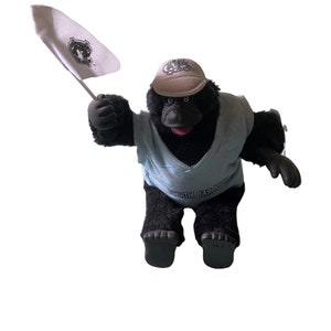 Op de afbeelding: Een zwarte pluche gorilla die een blauw shirt draagt met de tekst "NORTH CAROLINA". De gorilla draagt een beige baseballpet met een logo en houdt een witte vlag met een logo vast.