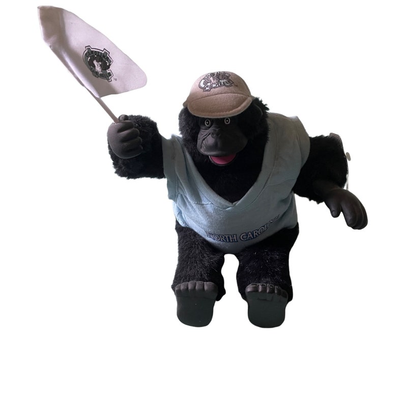 Op de afbeelding: Een zwarte pluche gorilla die een blauw shirt draagt met de tekst "NORTH CAROLINA". De gorilla draagt een beige baseballpet met een logo en houdt een witte vlag met een logo vast.