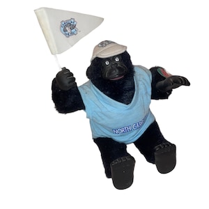 Op de afbeelding: Een zwarte pluche gorilla die een lichtblauw T-shirt draagt met "NORTH CAROLINA" erop. De gorilla draagt een witte baseballpet met "CAROLINA TAR HEELS" erop. De gorilla houdt een witte vlag vast met "CAROLINA TAR HEELS" erop.