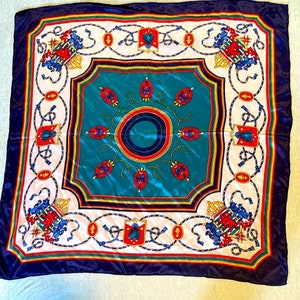 May include: A square silk scarf with a navy blue background and a white, red, and gold pattern. The pattern features a central circle with a blue and red design, surrounded by eight smaller circles with a similar design. The corners of the scarf are decorated with a repeating pattern of crowns and shields.