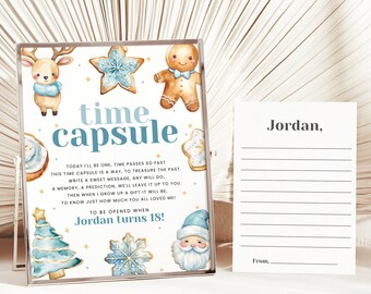Op de afbeelding: Een witte tijdcapsulekaart met een blauw en gouden winterthema. De kaart heeft een peperkoekman, een rendier en een sneeuwvlok. De tekst op de kaart luidt "time capsule" en "To be opened when Jordan turns 18!" Naast de tijdcapsulekaart ligt een blanco witte kaart met de tekst "Jordan," en "From..."