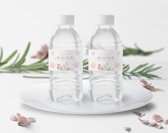 Peut inclure: Deux bouteilles d'eau en plastique transparent avec des étiquettes roses et blanches qui disent "Macy's Winter Wonderland". Les étiquettes représentent un cerf, un ours et des flocons de neige.
