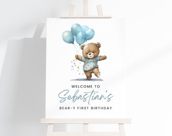 May include: Welcome to Sebastian's Bear-y First Birthday sign featuring a brown teddy bear holding blue balloons. The sign has a white background with blue text.