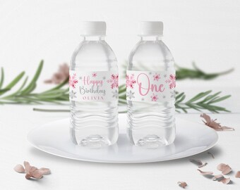Peut inclure: Deux bouteilles d'eau en plastique transparent avec des étiquettes personnalisées. Les étiquettes présentent un motif de flocons de neige rose et argent. Une étiquette dit "Joyeux anniversaire Olivia" et l'autre dit "Un".
