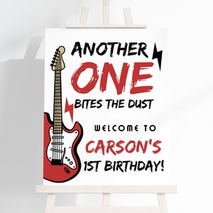 May include: A white sign with a red electric guitar and the text "Another One Bites The Dust" in black. Below the guitar is the text "Welcome to Carson's 1st Birthday!" in black.