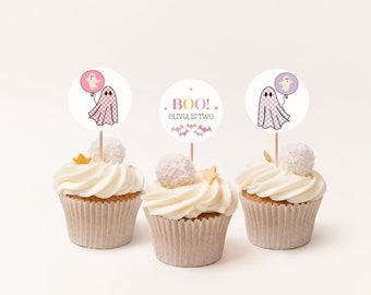 Op de afbeelding: Drie cupcakes versierd met witte frosting en bedekt met een witte chocoladebol. Elke cupcake heeft een ronde, witte papieren topper met een spookillustratie en de tekst "BOO! Olivia is two" in roze, geel en paars.