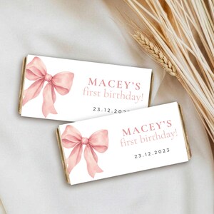 May include: Two rectangular chocolate bar wrappers featuring a watercolor illustration of a soft pink bow on the left side. The text reads MACEY'S first birthday! and the date 23.12.2023 in a simple, dark pink sans-serif font on a white background.
