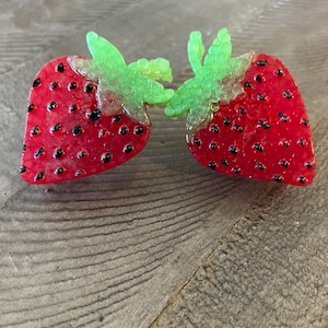 May include: Two red strawberry-shaped resin decorations with black seed details and green leafy tops. The strawberries are positioned close together, appearing to touch. The resin has a glossy finish.