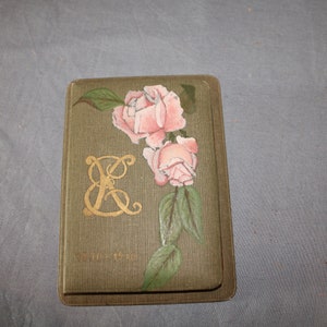 Può includere: Un libro verde con un monogramma in oro della lettera E e rose rosa dipinte sulla copertina. Il libro ha il testo "Aprile 18, 1917" stampato in basso.
