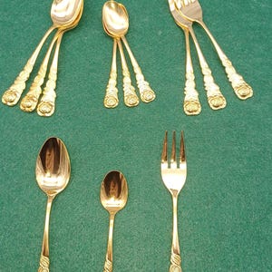 Può includere: Una collezione di posate color oro, tra cui cucchiai e forchette, disposte su una superficie verde. I manici presentano un design a rosa. Il set include cucchiai e forchette di varie dimensioni.