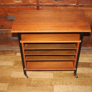 May include: A mid-century modern wooden and black metal shelving unit with four shelves and wheels. The unit has a top surface that can be used as a table.