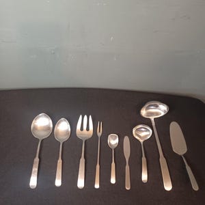 May include: A set of ten silver serving utensils, including a ladle, a serving spoon, a fork, a knife, and a spatula. The utensils are arranged in a row on a black surface.
