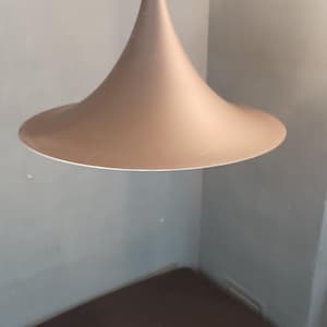 Könnte beinhalten: Eine braune, kegelförmige Pendelleuchte mit weißem Rand, die an einem dünnen Kabel aufgehängt ist. Das Licht hängt über einem dunkelbraunen Schrank vor einer gedämpften grünen Wand. Die Leuchte hat ein minimalistisches Design.