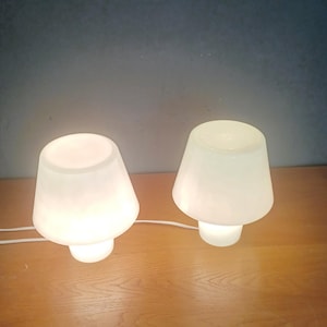 May include: Two white mushroom-shaped table lamps emitting a soft light. The lamps are made of opaque glass and sit on a wooden surface. Each lamp has a rounded base and a conical shade. The lamps are plugged in and lit.