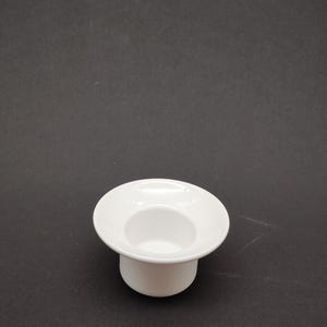 Könnte beinhalten: Ein weißer Eierbecher aus Keramik mit einem breiten, flachen Oberteil und einem abgerundeten Boden. Der Eierbecher ist für ein einzelnes Ei konzipiert und vor einem dunklen Hintergrund platziert. Das Design ist schlicht und modern.