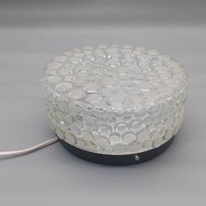 Könnte beinhalten: Eine Deckenleuchte aus klarem Glas mit einem strukturierten, blasenartigen Muster. Die Leuchte hat einen dunkelgrauen Sockel und ein weißes Netzkabel. Die Leuchte ist rund und scheint vintage zu sein.