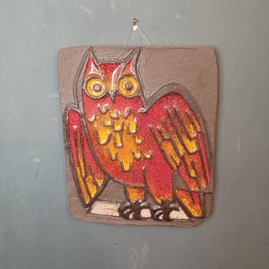 Könnte beinhalten: Eine Wanddekoration aus Keramik mit einem roten und gelben Eulenmotiv auf braunem Hintergrund.